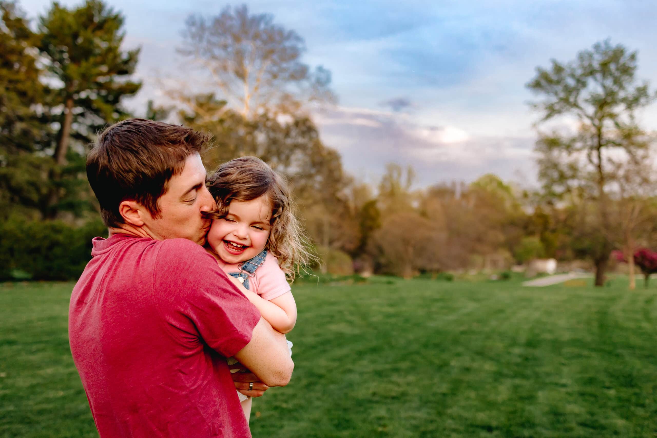 Dad hugging toddler in grassy field and giving her a kiss.