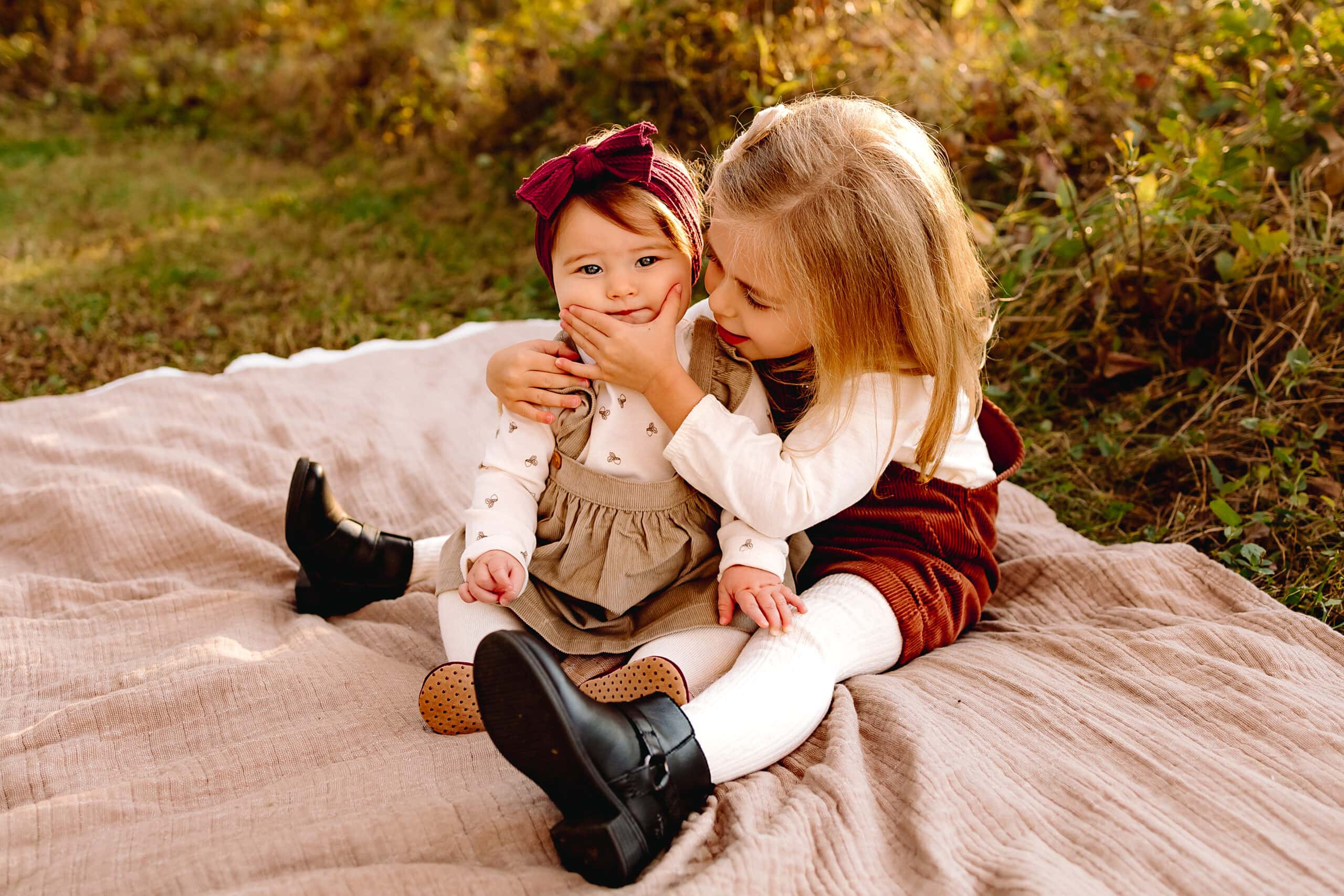 Sisters sitting on a blanket in a grassy field, the toddler is squishing the baby's cheeks.