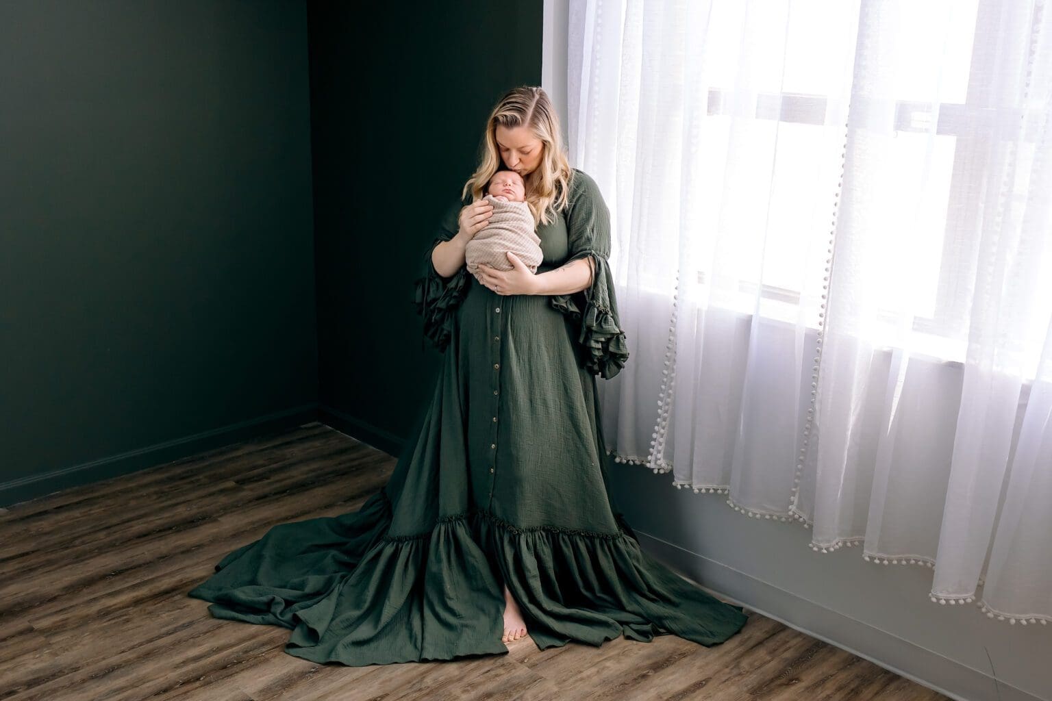 In a serene moment captured by the window's gentle light, a mother strikes a pose while holding her newborn wrapped in a soft tan wrap. With profound tenderness, she places a gentle kiss on the baby's head, expressing a deep affection and maternal adoration.