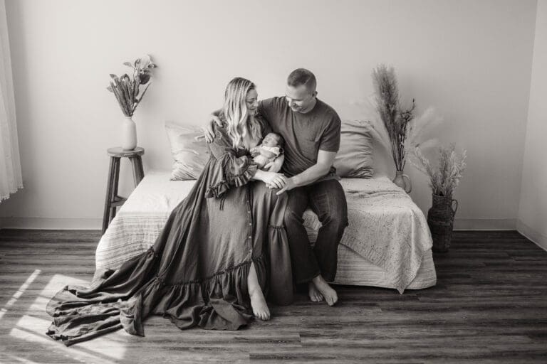 In a tranquil studio setting, a new mom and dad find solace on a comfortable daybed. The radiant mother cradles their precious newborn in her arms, while their eyes meet with love and tenderness. Their gentle interaction forms a captivating display of affection and a blossoming bond, capturing the joy and intimacy of parenthood.