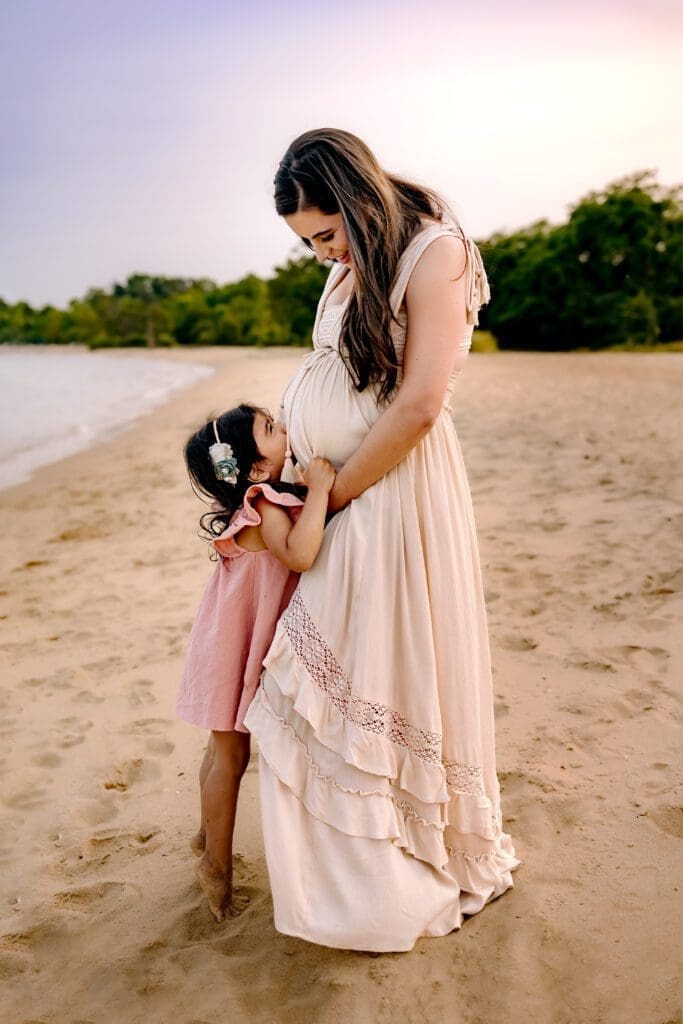Toddler daughter kisses mom's pregnant belly while having their summer maternity photos taken.