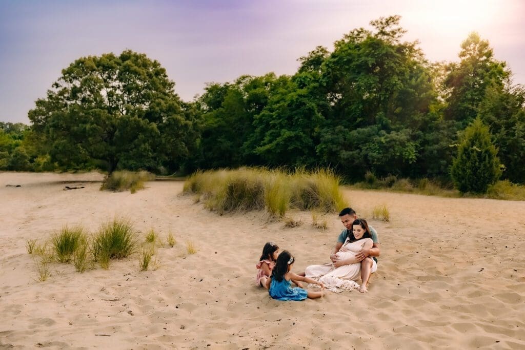 A family of four, with a glowing expectant mother, enjoy a relaxing moment on the beach during their photo session as they celebrate the impending arrival of their newest member.