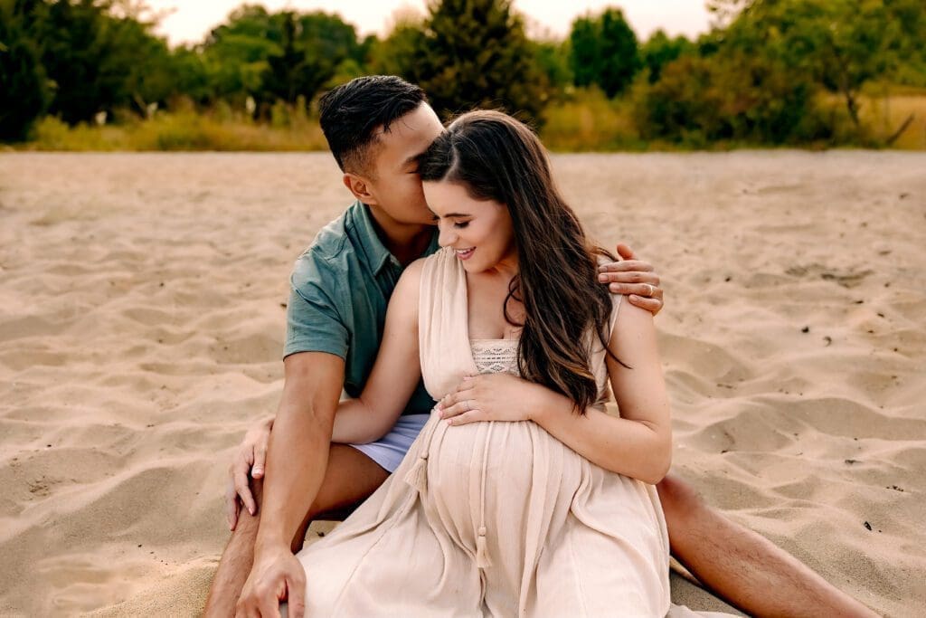 A tender moment captured as the expectant mother snuggles into her partner's arms, while he lovingly kisses her forehead during their beach maternity photoshoot.
