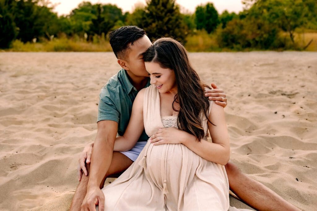 Crofton-Maternity-Photoshoot-8 A tender moment captured as the expectant mother snuggles into her partner's arms, while he lovingly kisses her forehead during their beach maternity photoshoot.