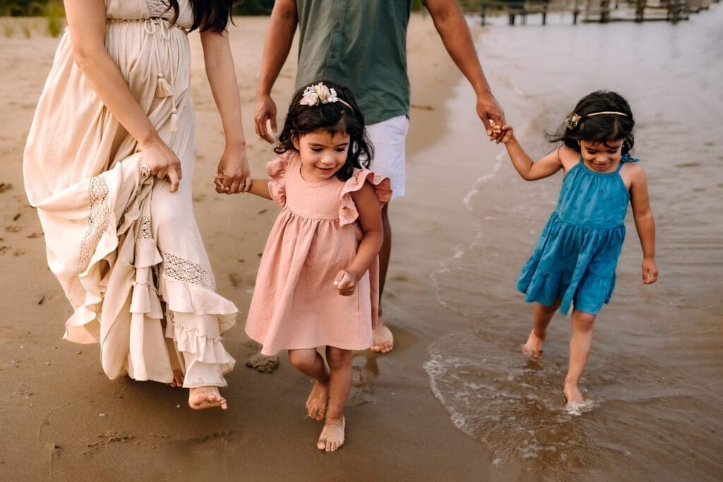 Mom and dad walk hand in hand, enjoying the shoreline as their two playful daughters splash in the waves during their summer maternity photo session.