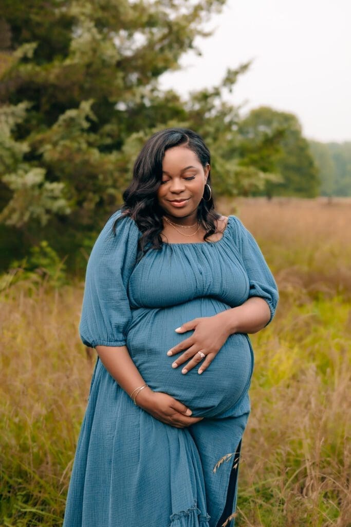 A mother holds her pregnant belly tenderly while standing in a lush green field in Piscataway Park, MD.