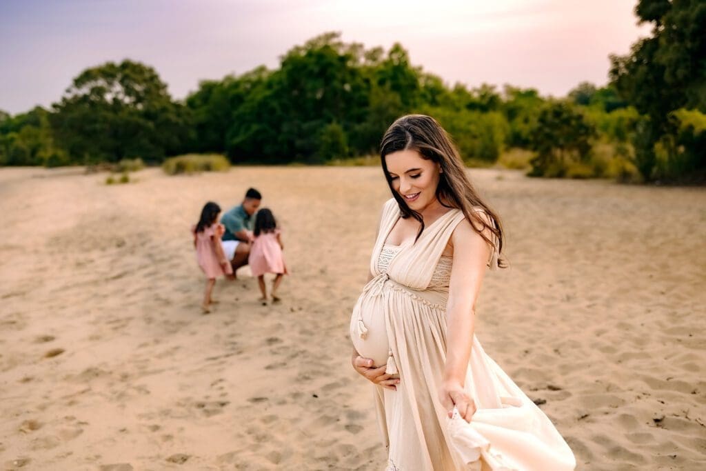 A joyful pregnant mom twirls her flowing dress on the beach for her summer maternity photos, while her husband and two adorable daughters play joyfully in the background.