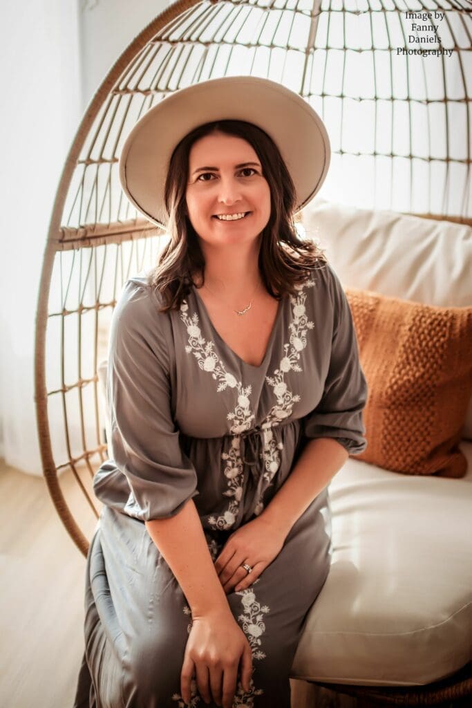 Erin Link, Annapolis Family photographer is sitting in an egg chair in a studio, holding a camera. She is wearing a blue embroidered dress and a hat, looking towards the camera with a smile. The studio is lit with natural light, creating a warm and inviting ambiance.