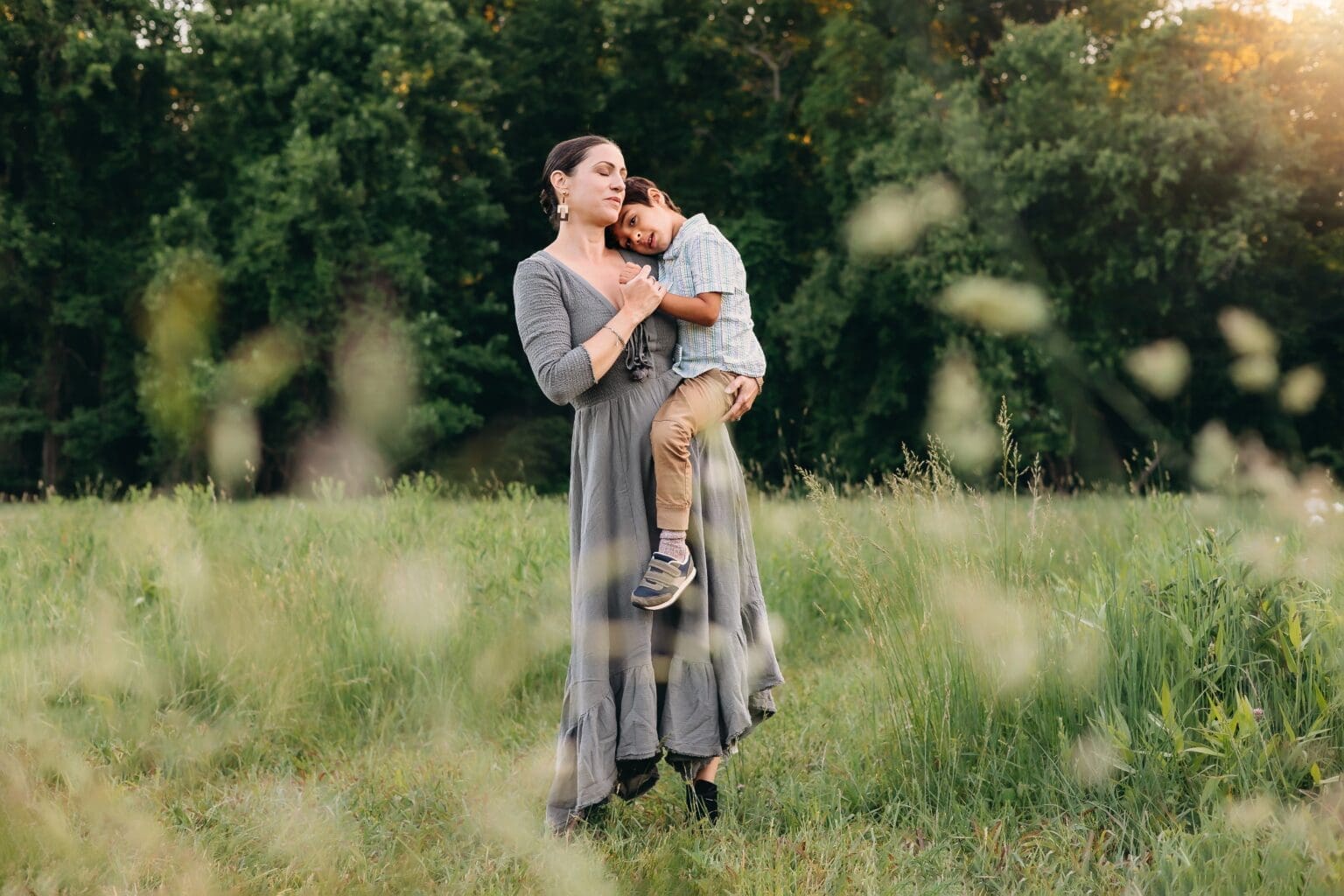 Mom holds her son as he rest his head on her shoulder in a lush green field.