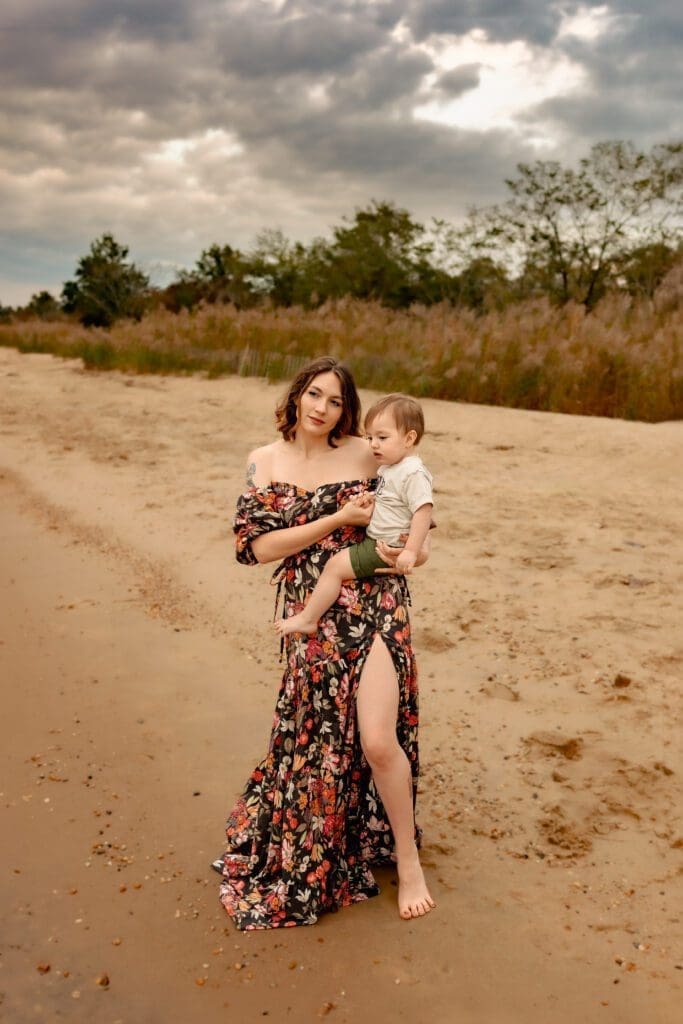 Mom wearing floral romance dress on beach in Maryland. Holds her baby on her hip and gazes out towards the water.