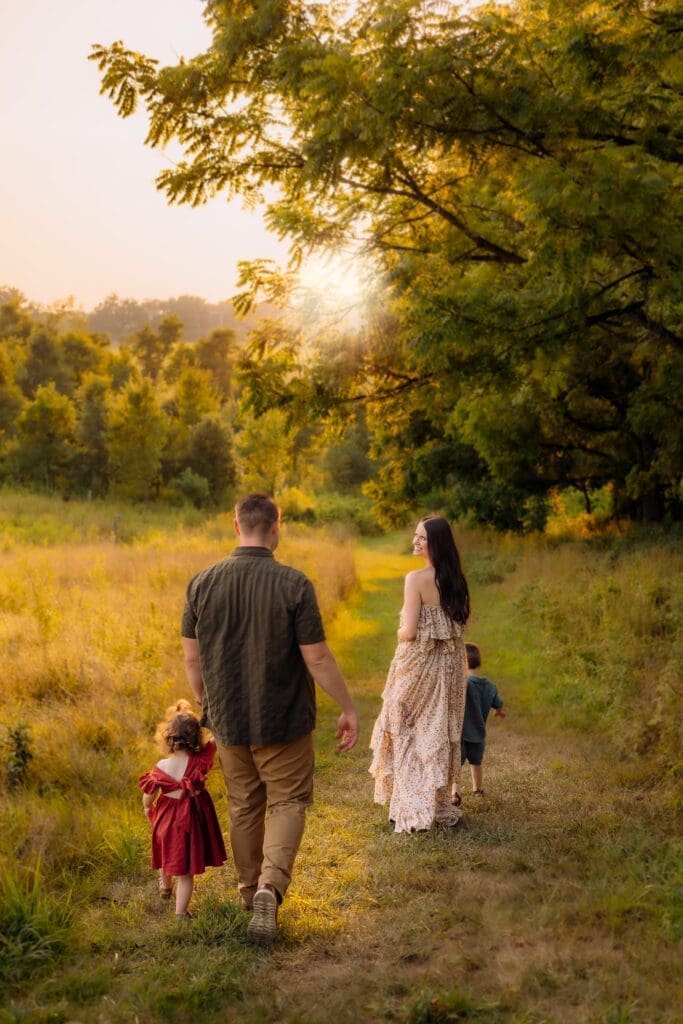 A happy family of four is taking a walk on a path at Howard County Conservancy. The mom is holding her son's hand and pausing to look back at her husband, who is walking with their daughter. The mom is smiling. The sun is shining and there are trees in the background, adding to the peaceful and serene atmosphere.