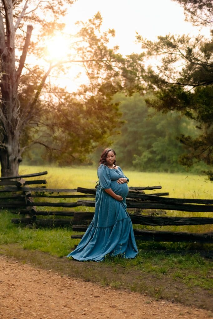 -A pregnant mother in a teal dress is posing in Piscataway Park in Maryland. She is lovingly holding her belly while looking off over her shoulder. The sun is shining in the background, and the trees are lush and green.