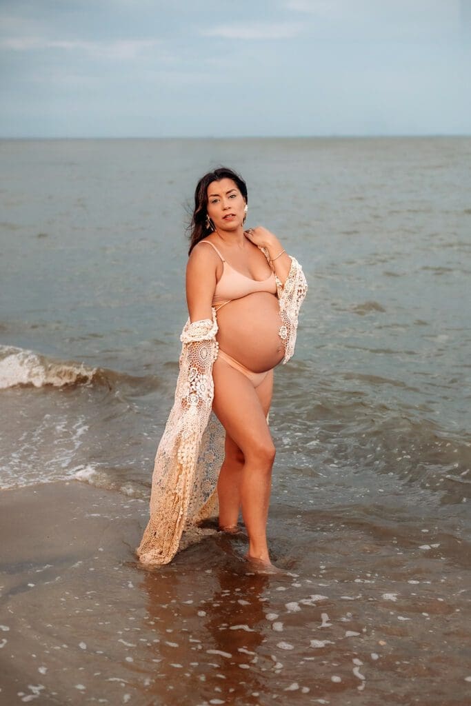 Pregnant mom in crochet robe on beach. She gazes into the camera as she walks along the water.