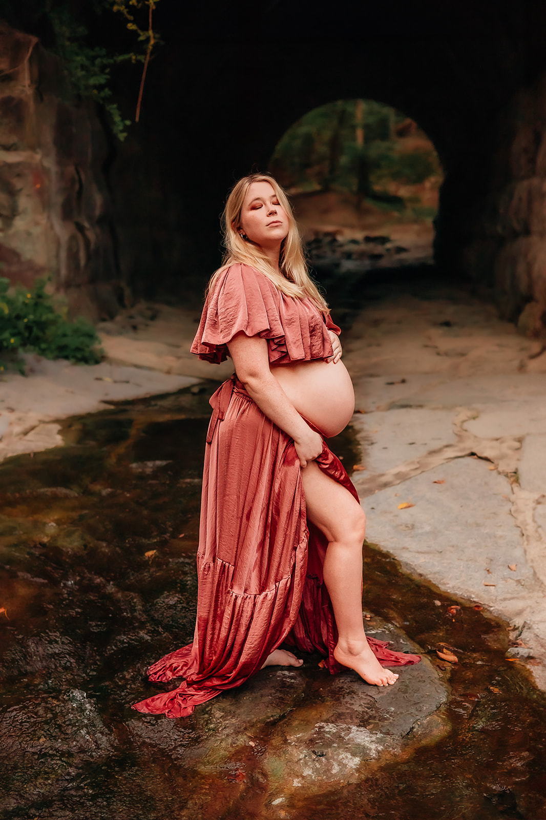 Pregnant mom in a mauve dress poses gracefully in front of a tunnel. Her eyes are closed, and she tilts her head upward, creating a serene and contemplative moment. The soft glow of the tunnel's light enhances the beauty of the scene, capturing the anticipation and joy of pregnancy.