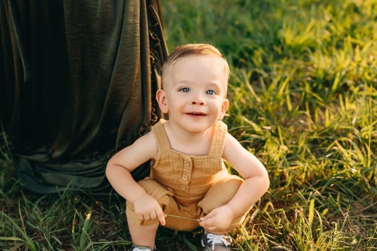 Sun-kissed smiles light up the frame during toddler photo sessions. Toddler basks in the golden glow in a field at Kinder Farm Park in Maryland.