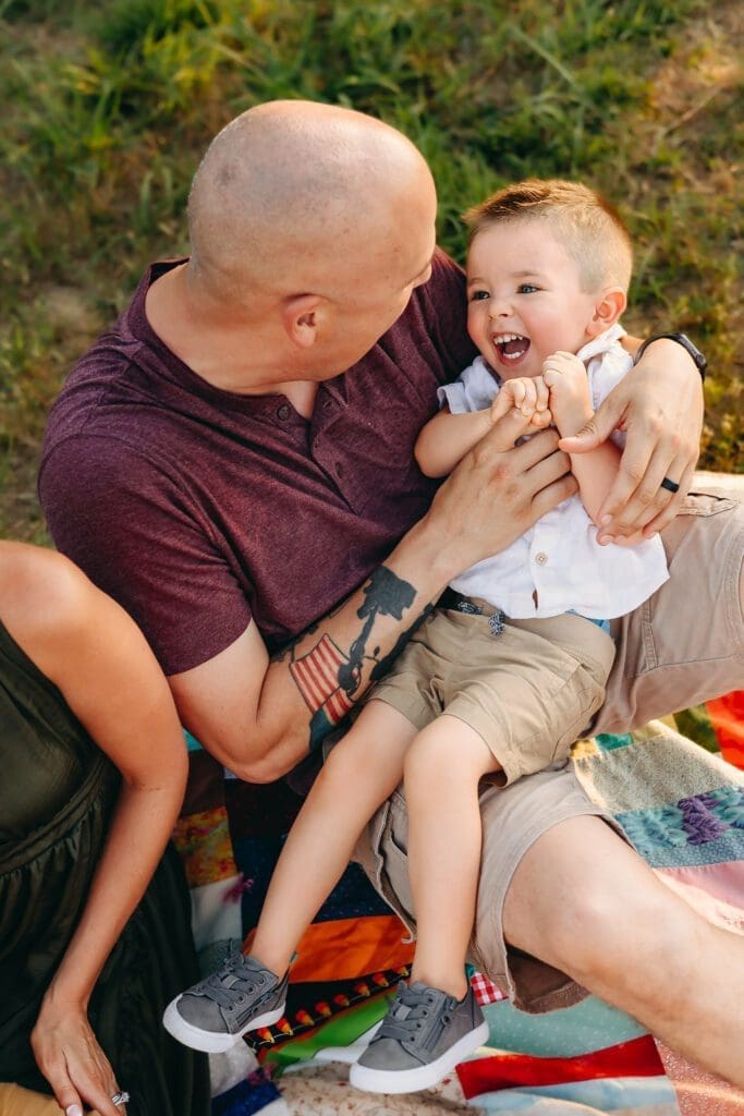 Dad holds his toddler on his lap and gives him a tickle. They sit on the grass in a field in Kinder Farm Park.