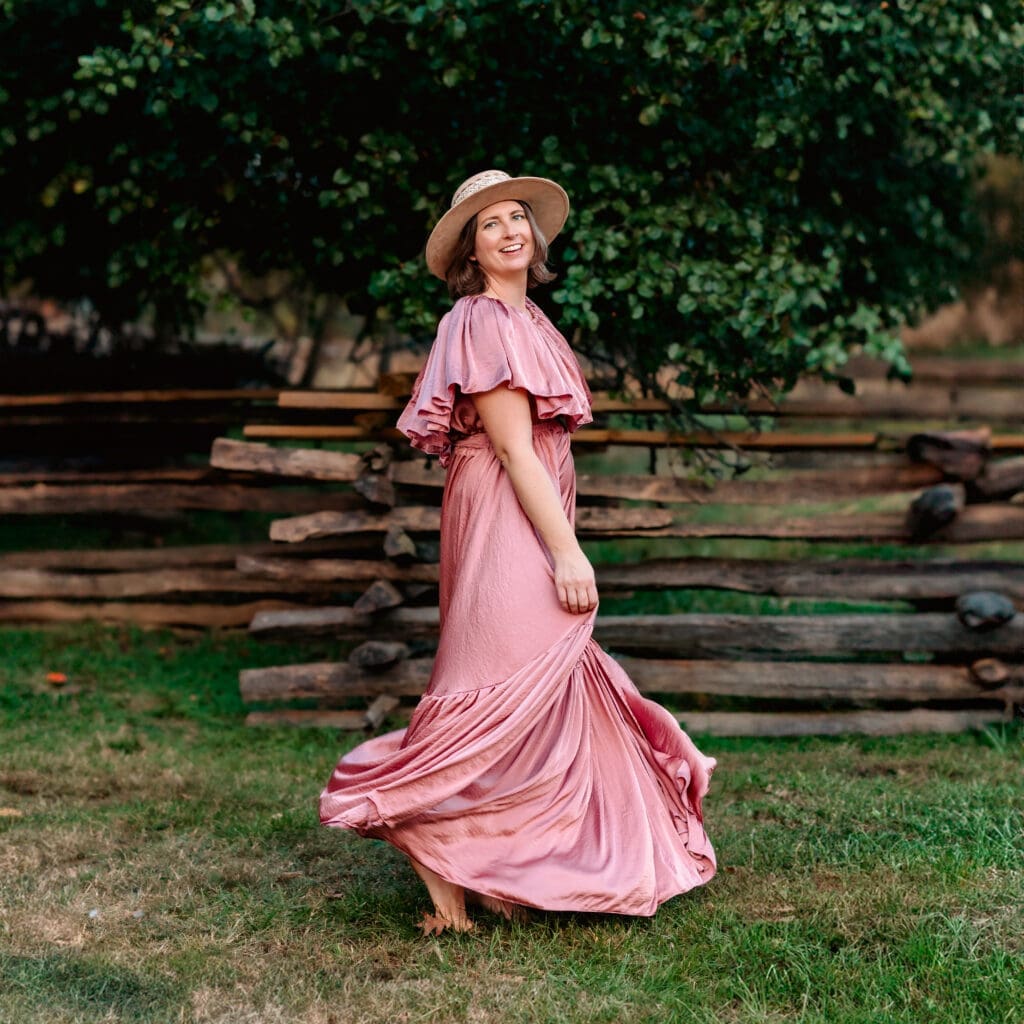 Portrait of Erin Link, a smiling family photographer in a flowing pink dress and a suede hat, standing in a pastoral setting with lush trees and a rustic wooden fence in the background, conveying a sense of warmth and ease in nature.