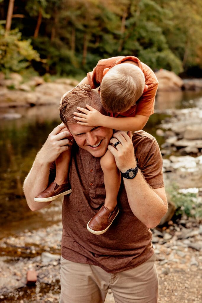 Father and son sharing a playful piggyback moment by the river, with genuine smiles and laughter, perfect for a family lifestyle photography session.