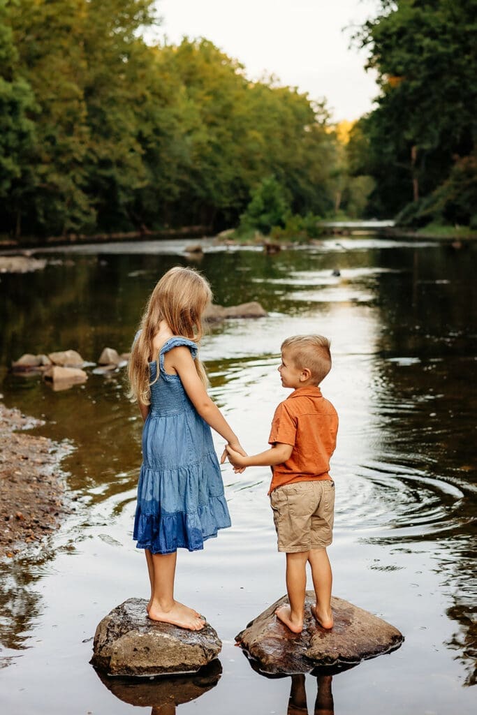 Two young siblings holding hands on stepping stones in a river, an idyllic scene for a family lifestyle photography session