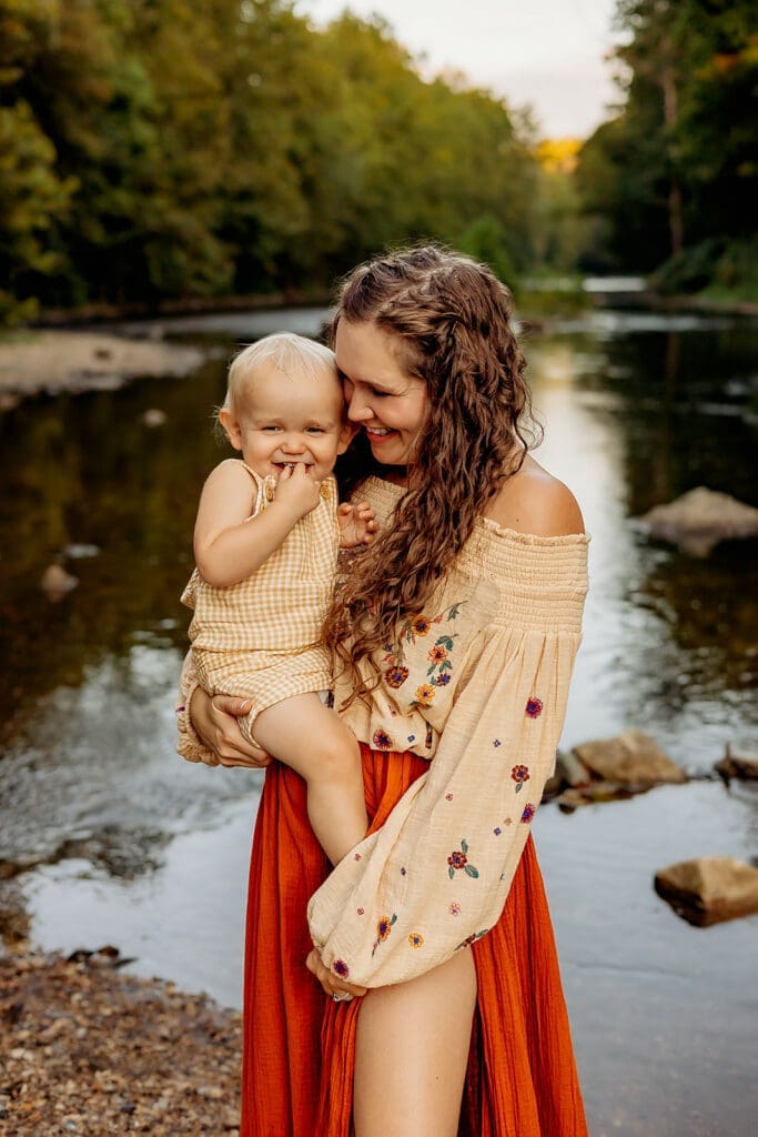 Mother holding her toddler, both smiling warmly, against a backdrop of a serene river, capturing the essence of a joyful family lifestyle portrait.