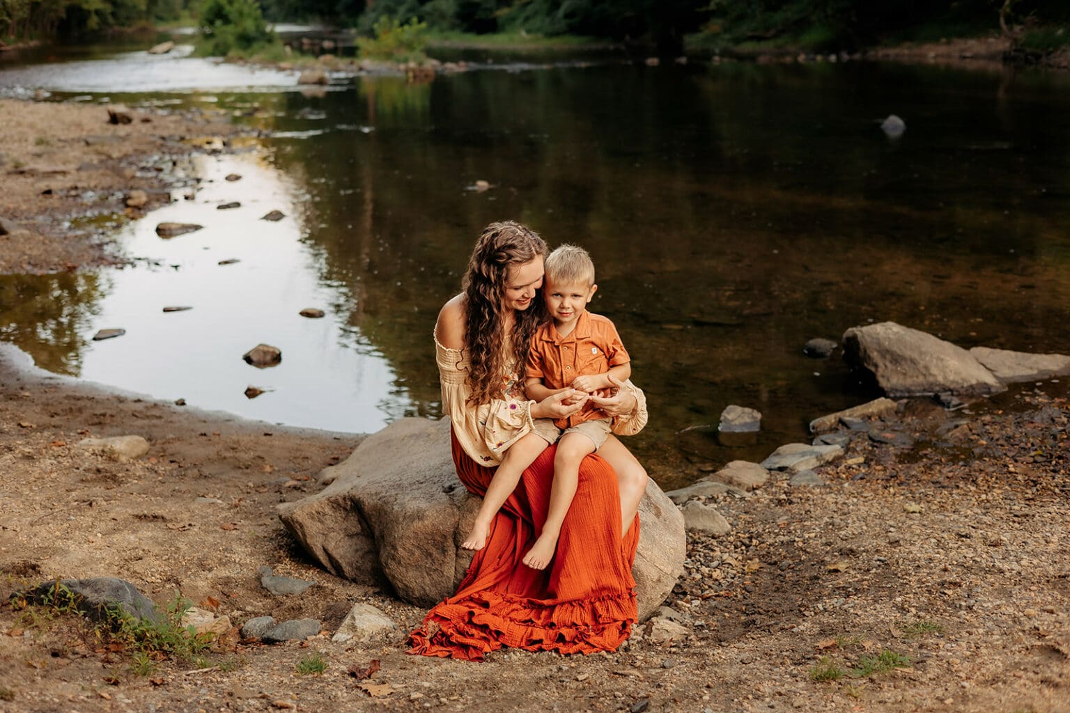 Young mother and son sitting on a rock by a tranquil river, sharing a moment of closeness in a picturesque outdoor lifestyle photography session.