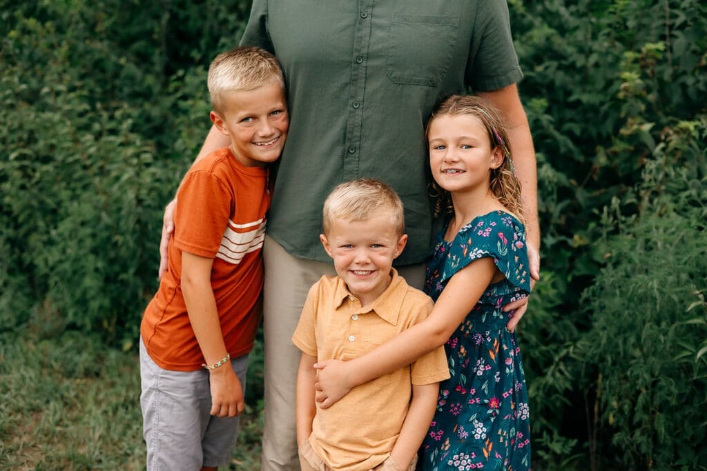 The family stands closely, the father embracing the two sons and daughter, all kids are smiling at the camera