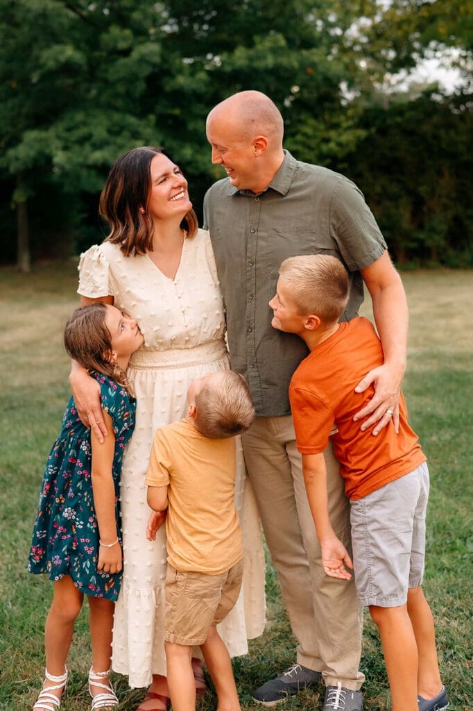 The family stands closely together looking at each other; the parents are in the center with the children around them.