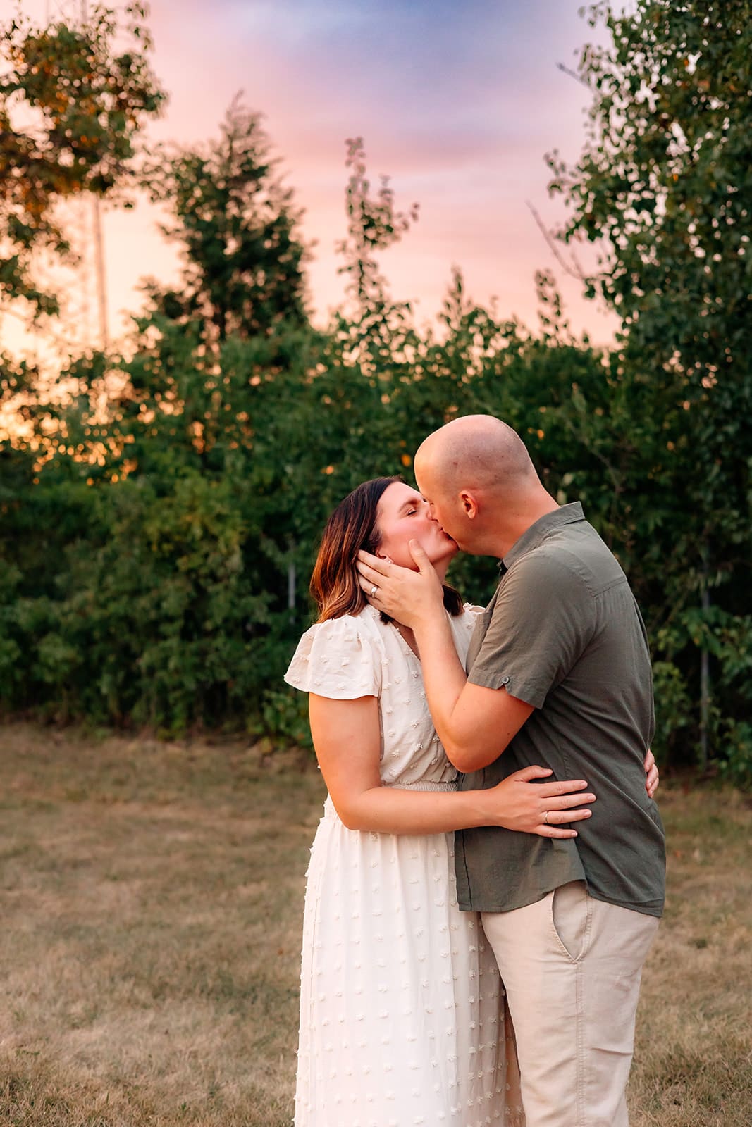 Parents are embracing and kissing, with a backdrop of trees and a twilight sky.
