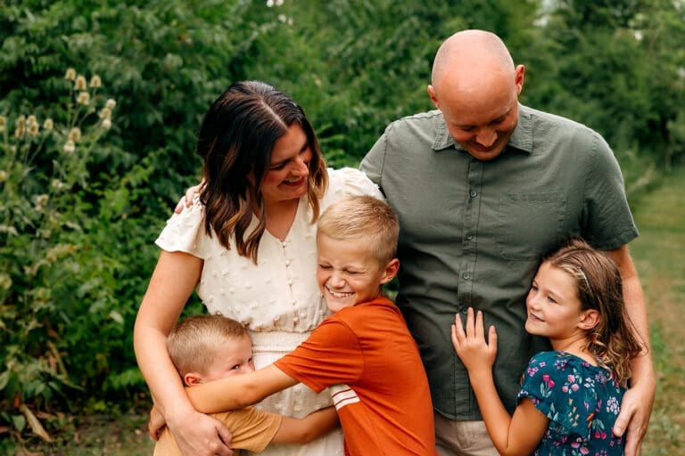 A family of five with two boys and a girl, the parents are looking at their daughter while the boys hug the mother's legs.