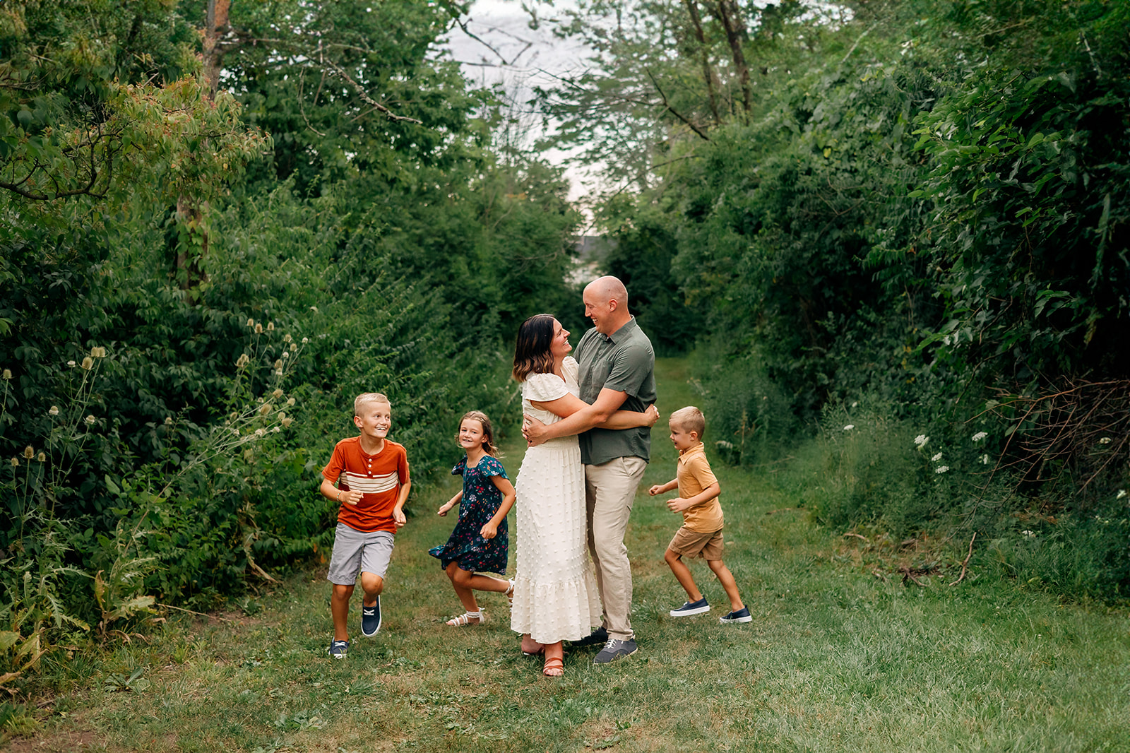 The image shows a candid moment of a family outdoors. In the center, a couple is embracing, the woman leaning into the man as they both smile softly. To their left, two children, a boy in an orange shirt and a girl in a navy floral dress, are laughing and running towards the camera. On the right, another boy in a tan shirt smiles as he looks ahead, running slightly ahead of the rest. The background is lush with greenery, indicating a natural, possibly wooded area. The scene is lively and portrays a sense of movement and familial joy.