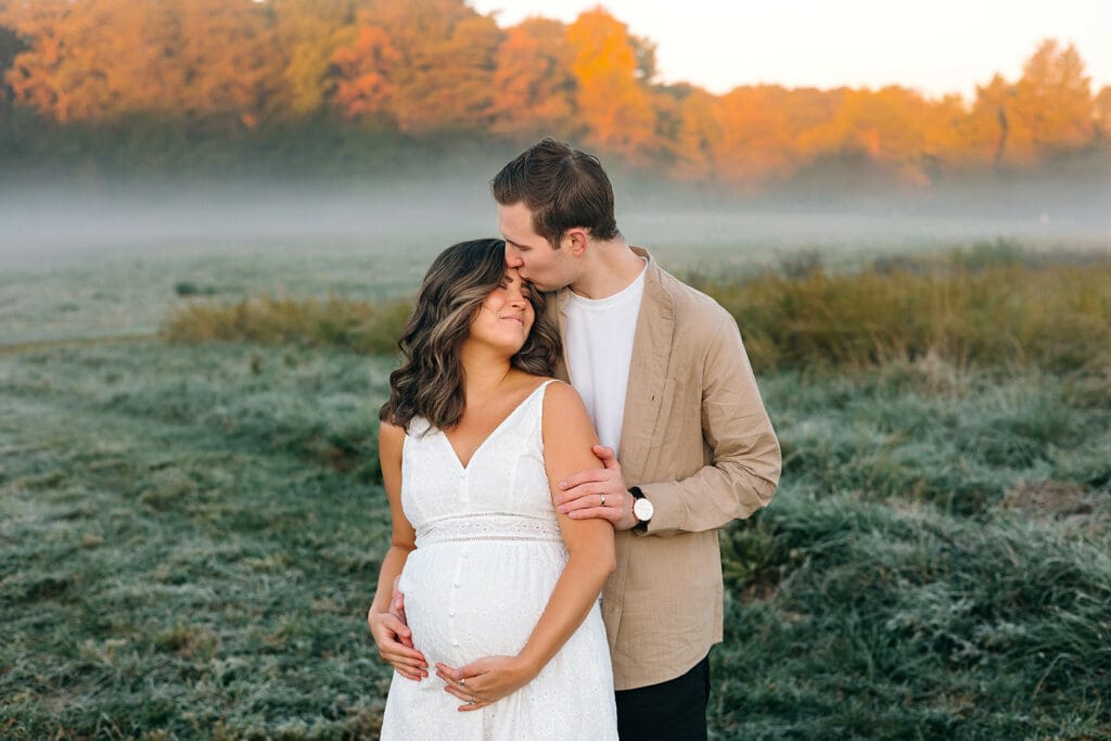 Couple in a maternity photoshoot embracing amidst morning fog in an autumn field.
