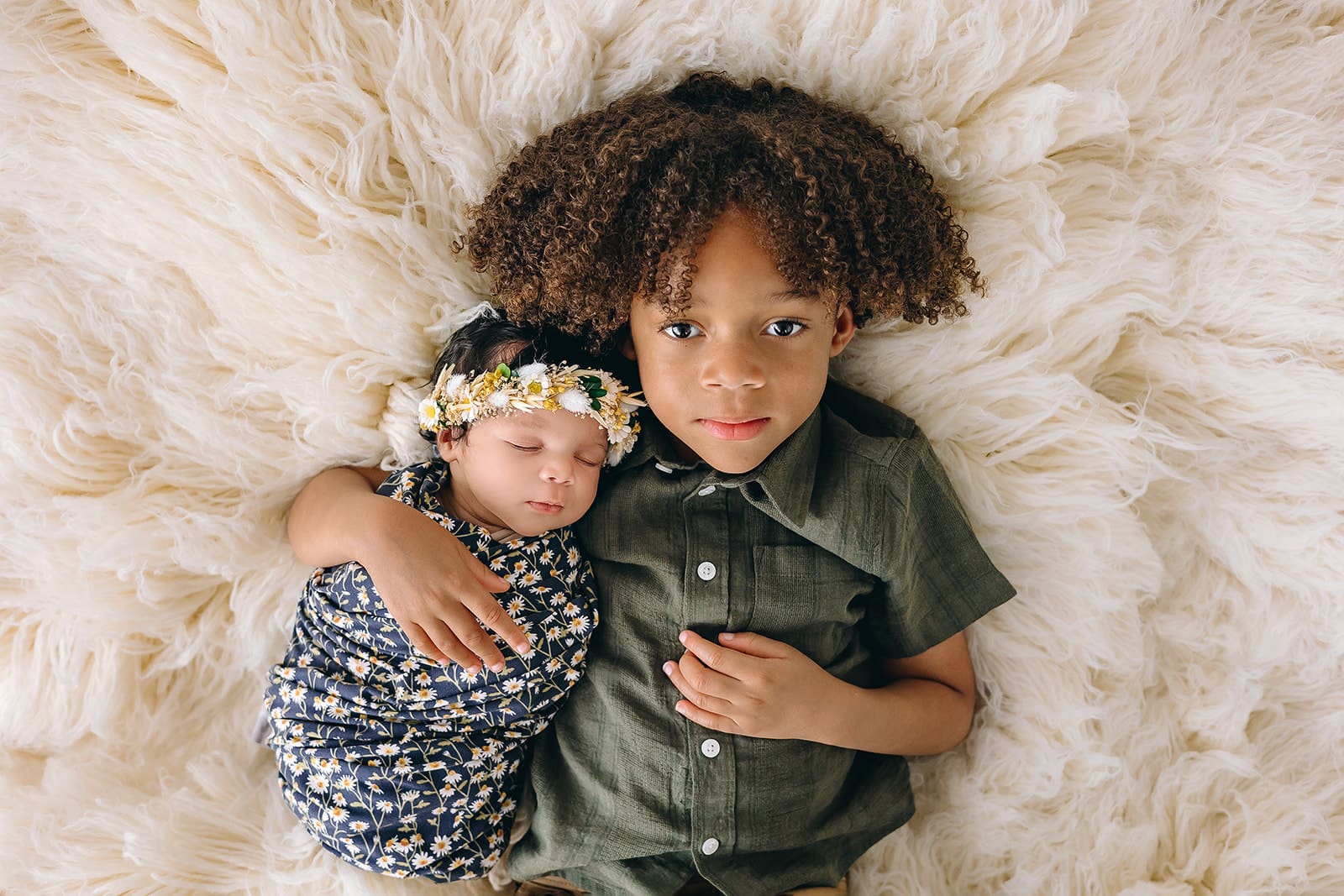 An adorable top-down view of two siblings lying on a fluffy white rug. The newborn, wrapped in a navy floral swaddle and wearing a delicate floral headband, sleeps peacefully. Next to the baby is the older sibling, a curly-haired child wearing a buttoned olive-green shirt, gazing at the camera with an expression of pride and happiness.
