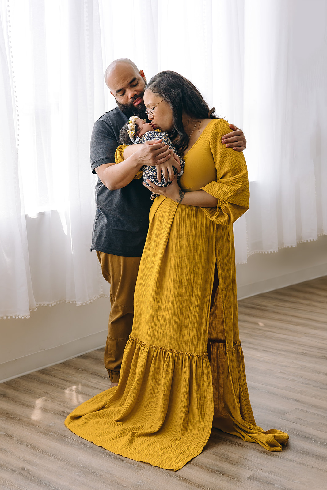 A heartwarming portrait of a family in a cozy studio setting. The father, with a closely shaved head and beard, dressed in a grey T-shirt and mustard trousers, tenderly embraces his partner and their newborn. The mother, wearing glasses, a long mustard dress with puffed sleeves, cradles the swaddled baby lovingly in her arms. She is standing and smiling down at the infant. A sense of peaceful joy permeates the room with sheer curtains softly diffusing the daylight.