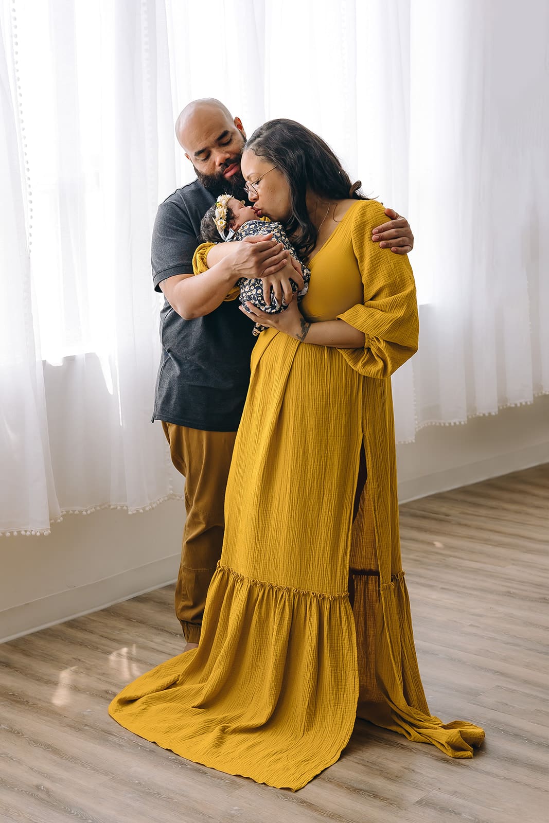 Lifestyle Studio Newborn Session A heartwarming portrait of a family in a cozy studio setting. The father, with a closely shaved head and beard, dressed in a grey T-shirt and mustard trousers, tenderly embraces his partner and their newborn. The mother, wearing glasses, a long mustard dress with puffed sleeves, cradles the swaddled baby lovingly in her arms. She is standing and smiling down at the infant. A sense of peaceful joy permeates the room with sheer curtains softly diffusing the daylight.