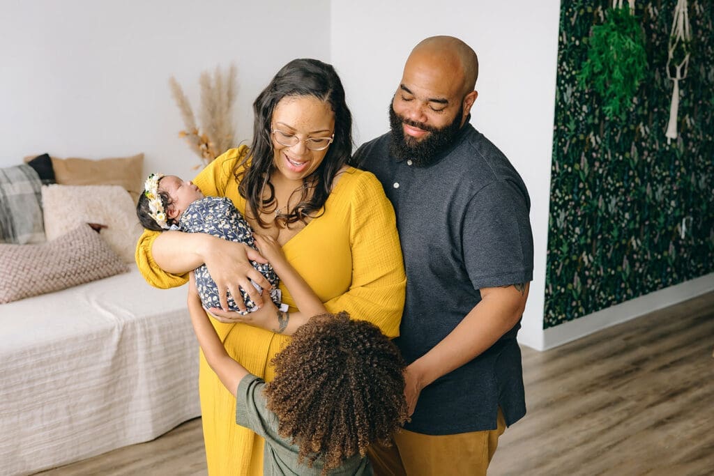 A candid moment captured in a lifestyle studio, featuring a family of four. The mother, dressed in a mustard yellow dress, is laughing joyously as she holds her swaddled baby close, the infant adorned with a cute floral headband. The father, in a grey T-shirt and black shorts, stands behind them, smiling contentedly. Their older child, in an olive-green shirt, stands in front, looking up at them with a sense of inclusion and love.