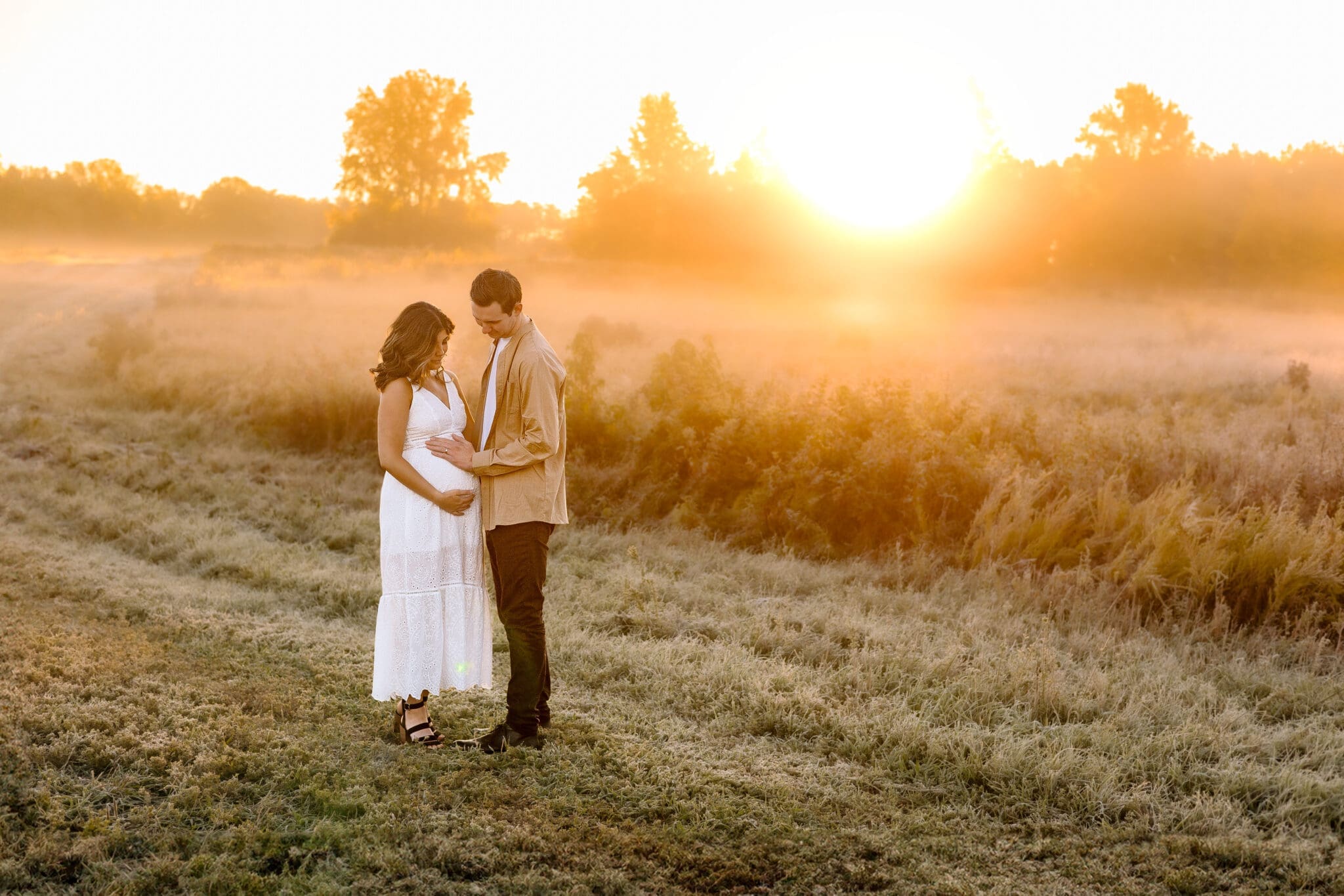 Couple standing in a field with a sunrise backdrop during a maternity photoshoot.