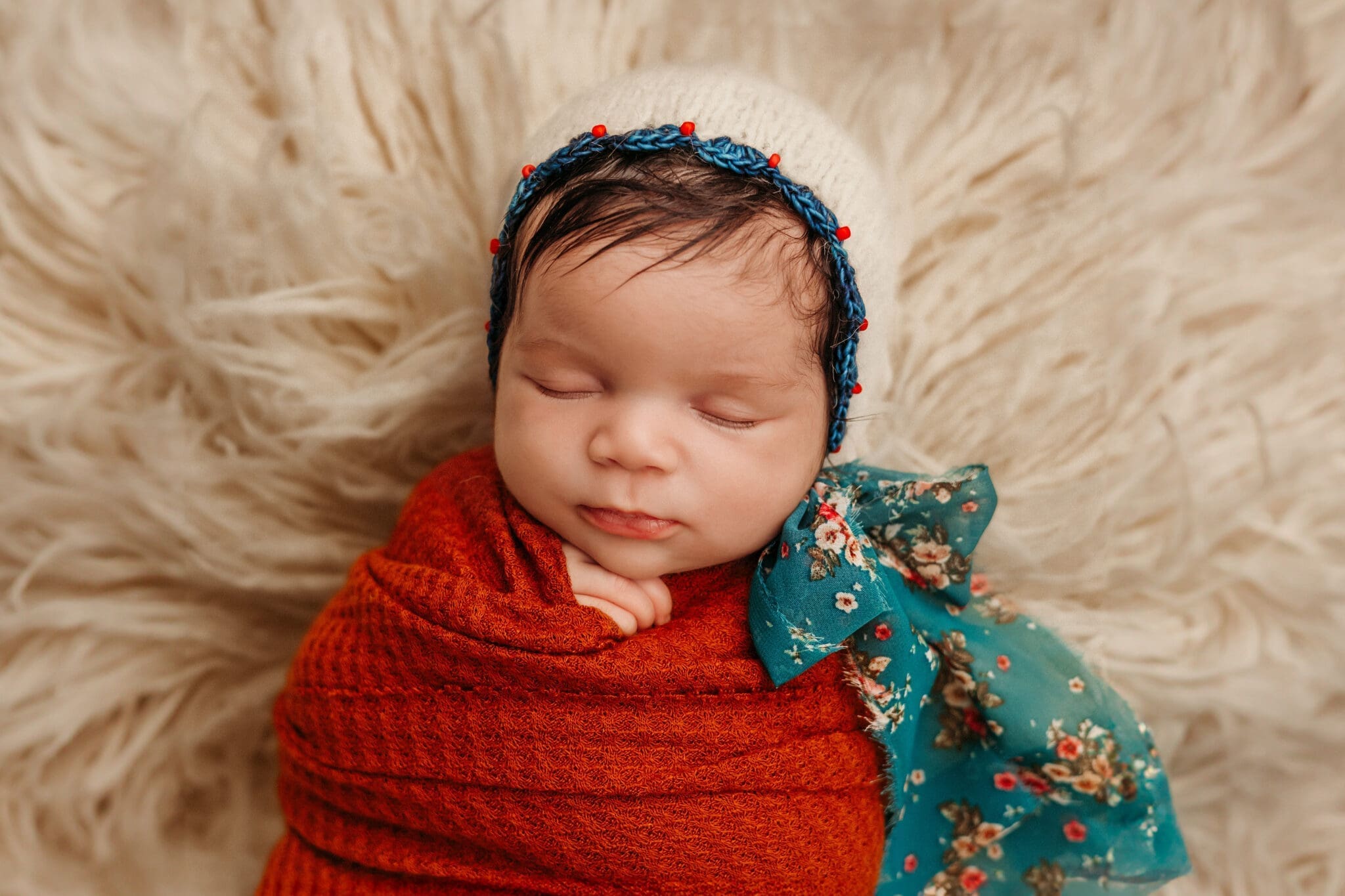 A serene close-up of a sleeping newborn swaddled in a rich rust-colored blanket, lying on a fluffy cream-textured rug. The baby is wearing a white knitted bonnet. The infant's delicate features are relaxed in sleep, and there's a faint hint of a smile on their lips.