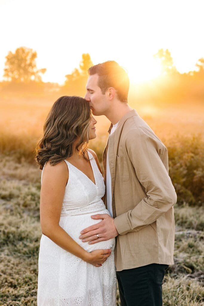 Couple holding each other in a warm embrace in a foggy meadow. Dad lovingly kisses mom on the forehead.