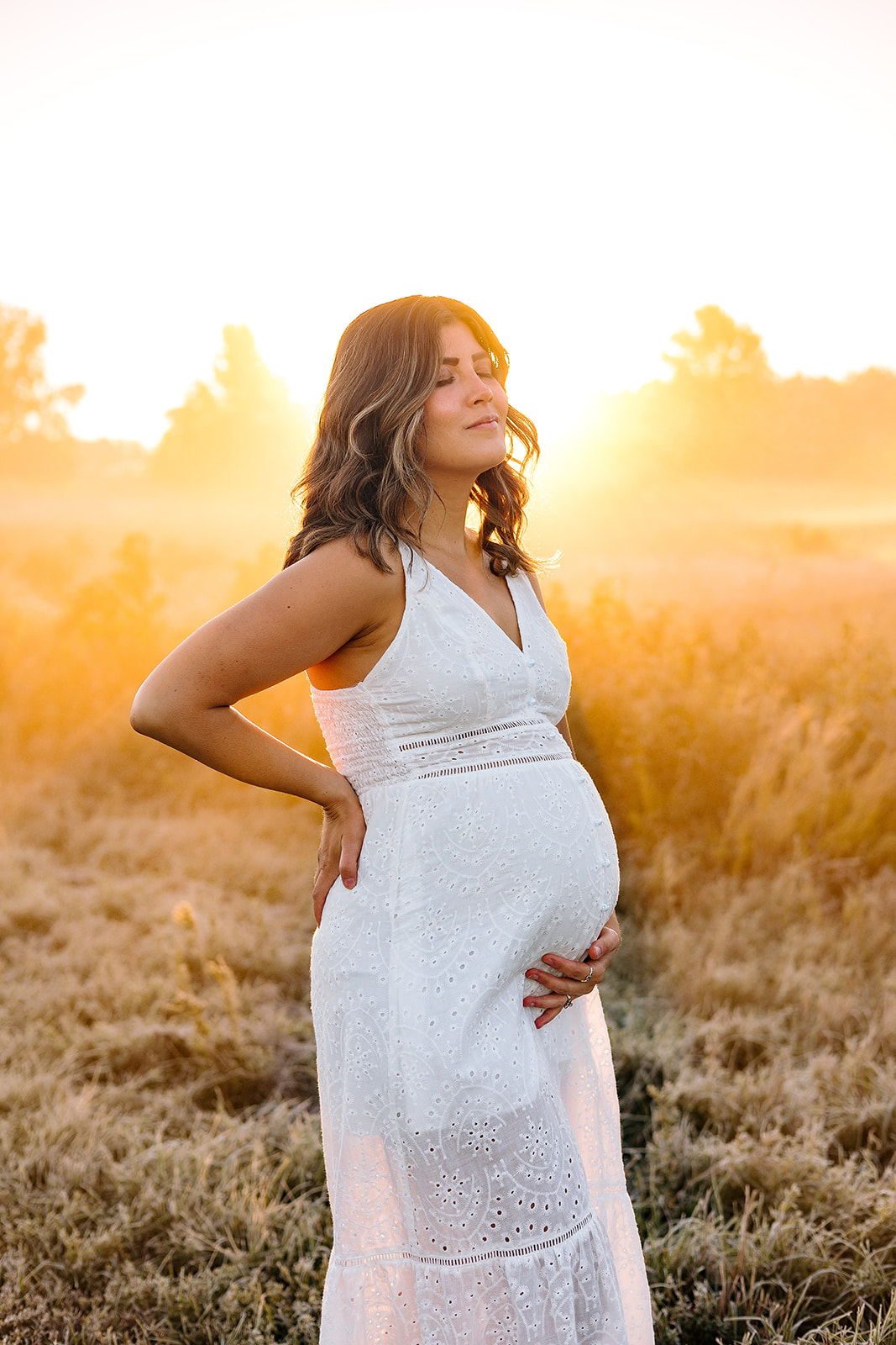 Maternity photoshoot tips featuring a woman in a white dress cradling her belly, standing alone in a field.