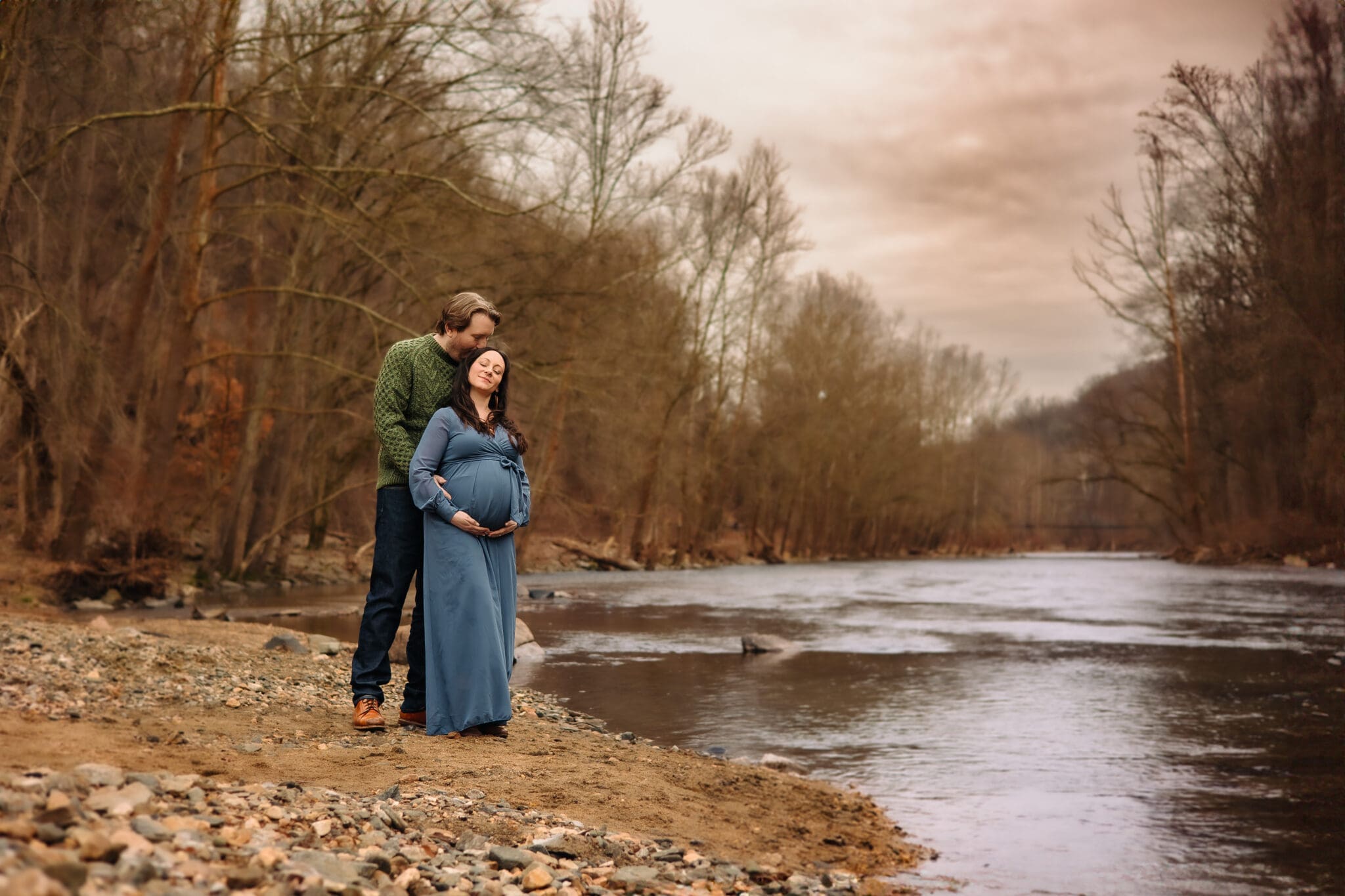 Pregnant woman in a long blue dress standing by a peaceful river, embraced by her partner in a green sweater. Bare winter trees line the background, creating a serene and intimate moment.