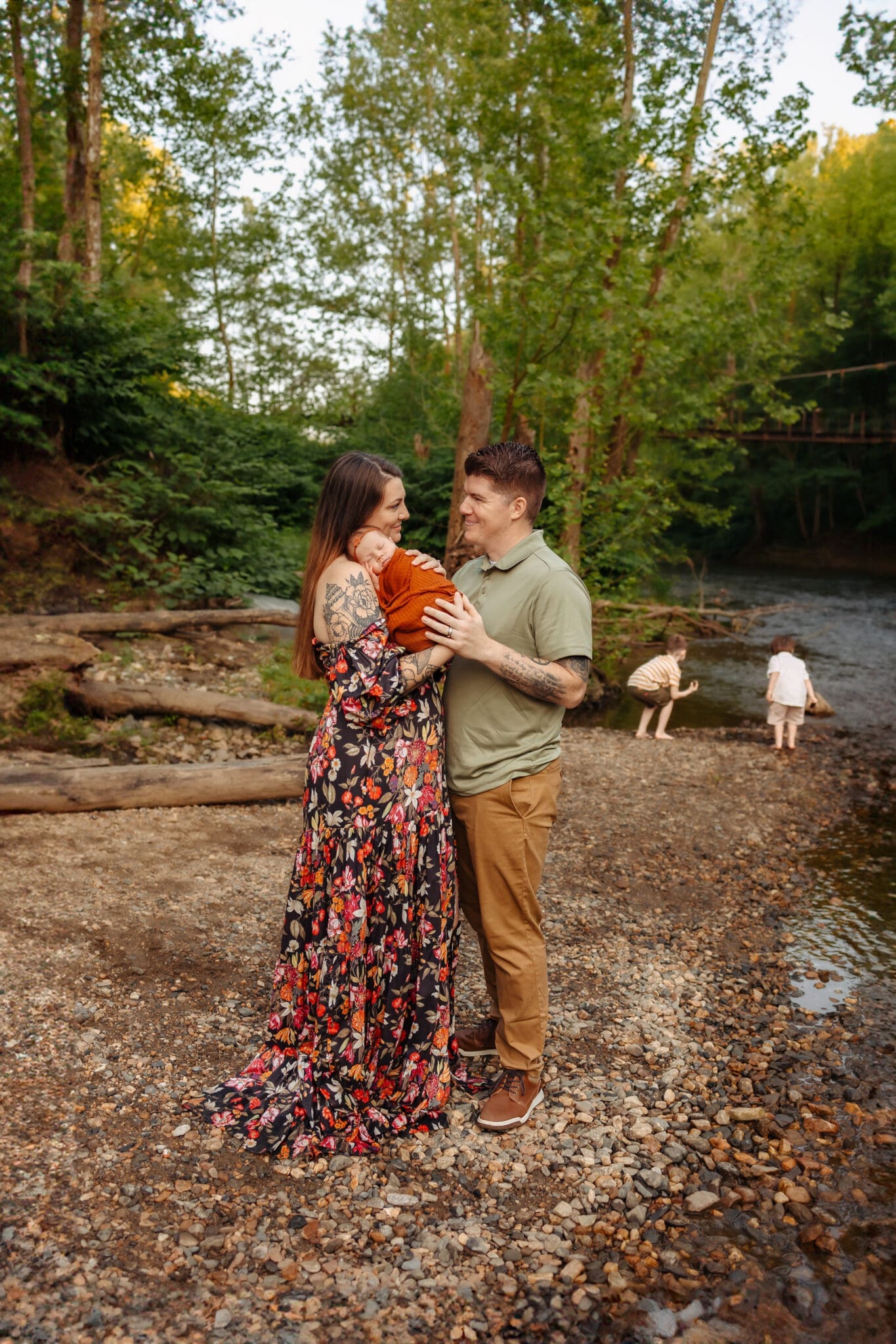 A mother and father standing close together, holding their newborn baby wrapped in a rust-colored swaddle by the river. Photo by Erin Link Photography.