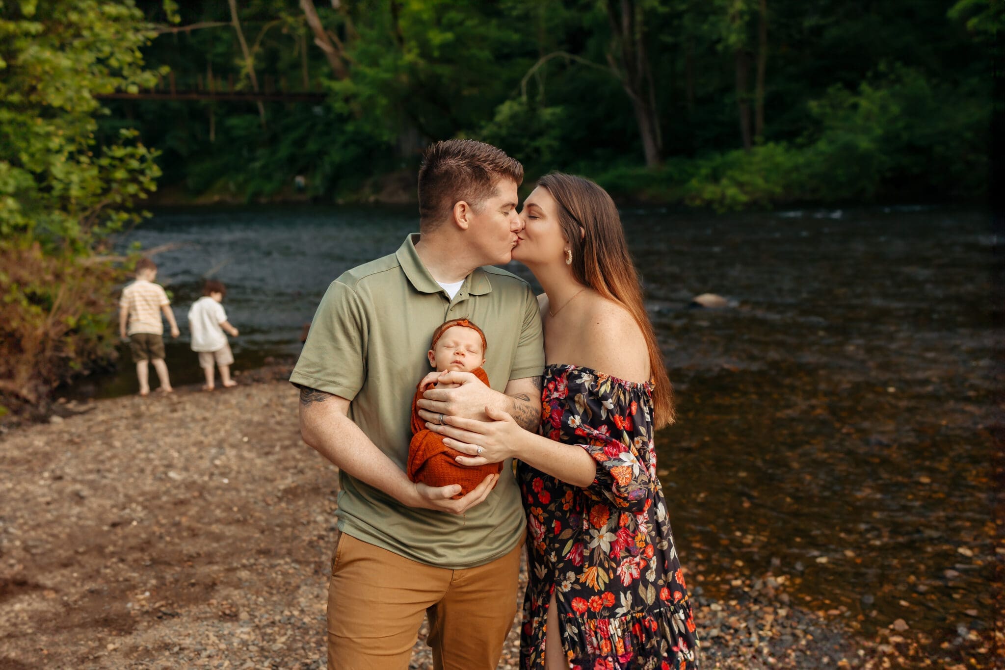 newborn-photography-erin-link A couple after preparing for baby in Bowie, stand kissing while the father holds their newborn baby. Two older children are playing in the background near a river."