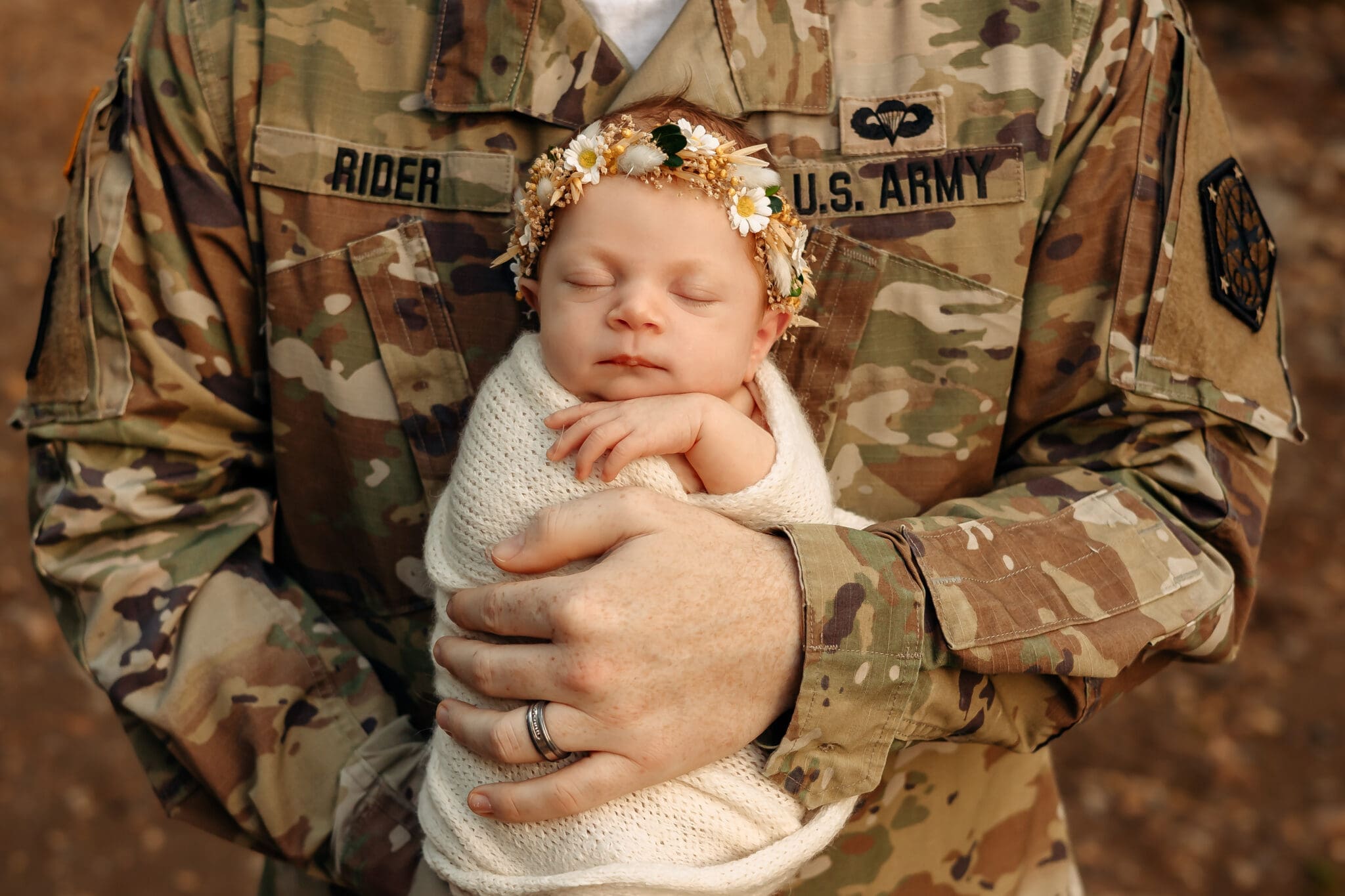 A U.S. Army soldier in uniform holding a newborn baby wrapped in a cream blanket with a floral headband.