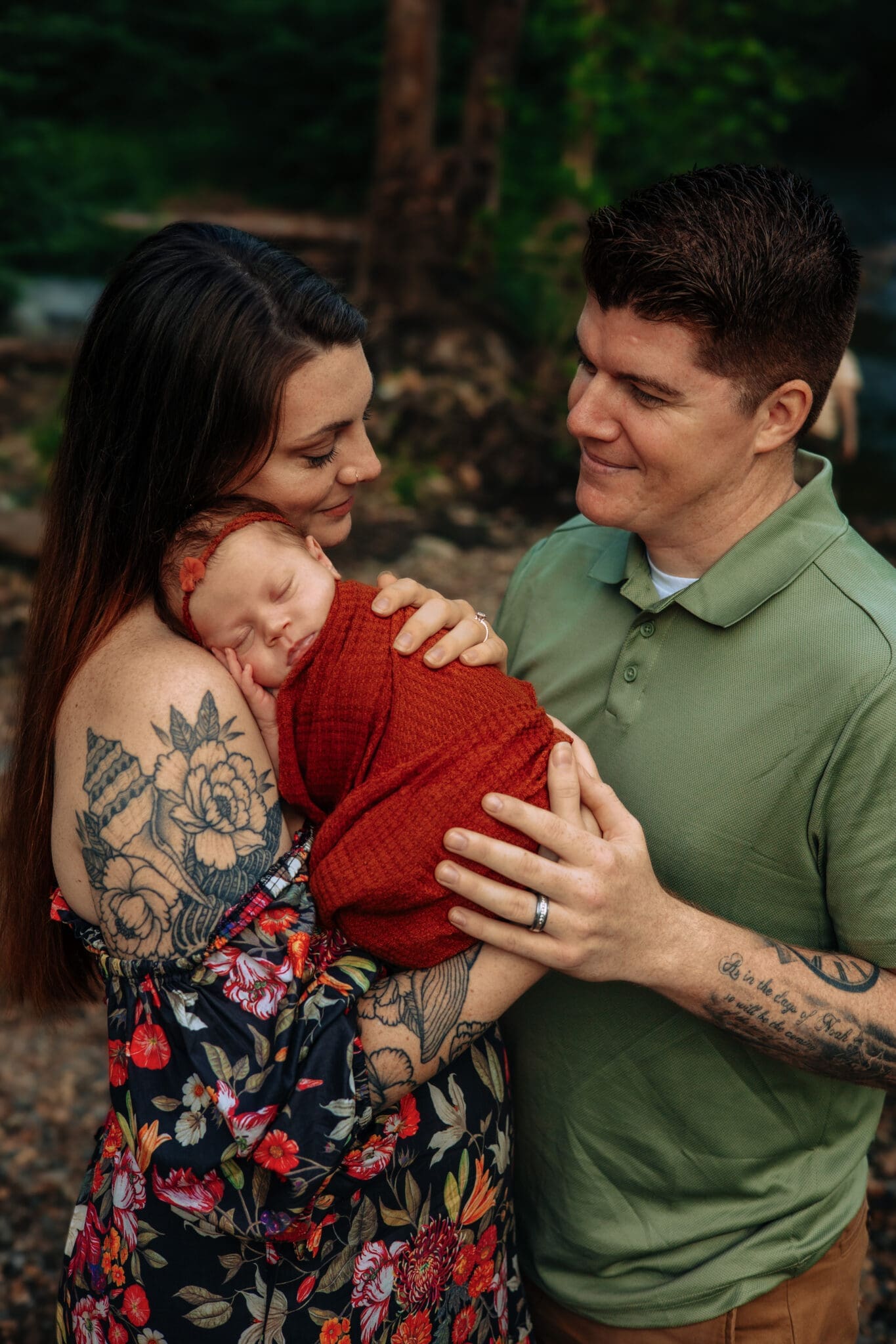 A tender newborn family moment, with a sleeping baby wrapped in a rust-colored blanket, held close by loving parents during an outdoor session in Maryland.