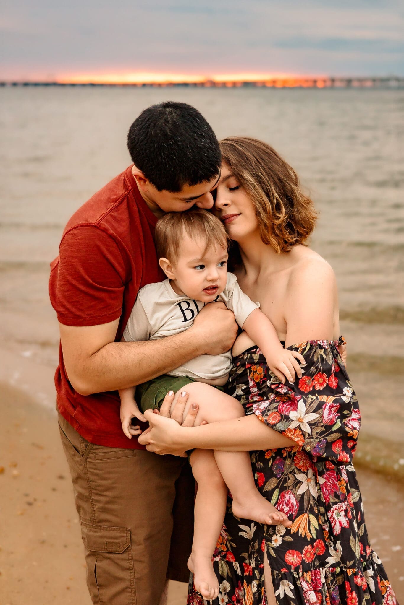 A young family embraces on the shore during sunset. The mother, in an off-the-shoulder floral dress, holds her barefoot toddler, while the father, in a red shirt, wraps them both in a hug, kissing his child's head. The soft golden light reflects off the water, creating a warm and intimate moment.