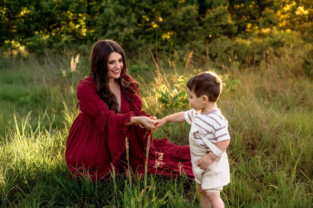 A pregnant mother in a flowing red dress kneels in a sunlit field, holding hands with her young son during their spring maternity photos in Annapolis.