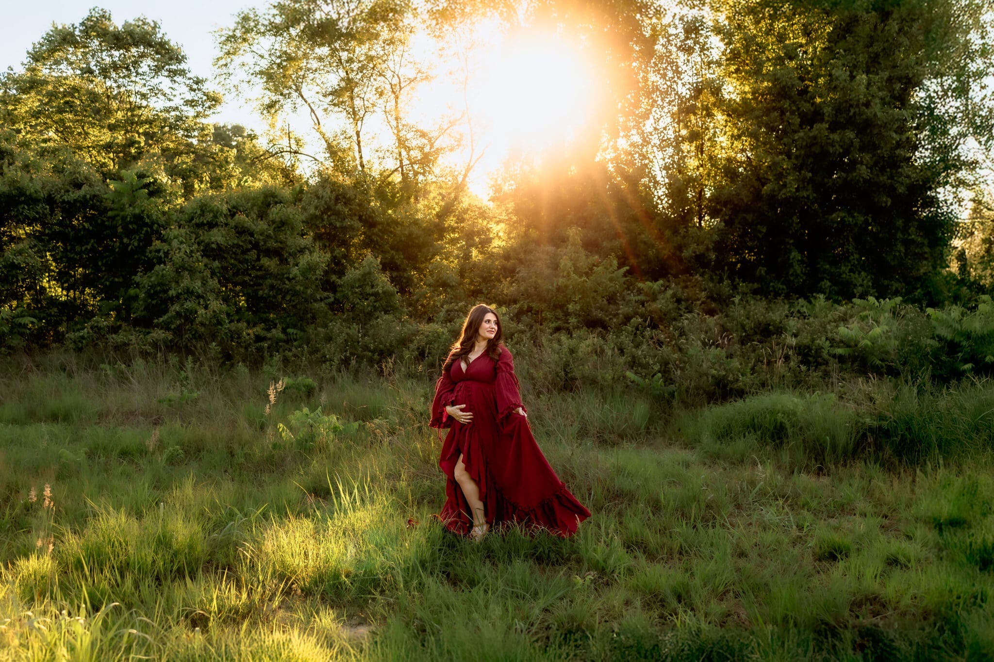 A mother-to-be in a flowing red dress walks through a green meadow at sunset, surrounded by nature, during her spring maternity photos in Annapolis.