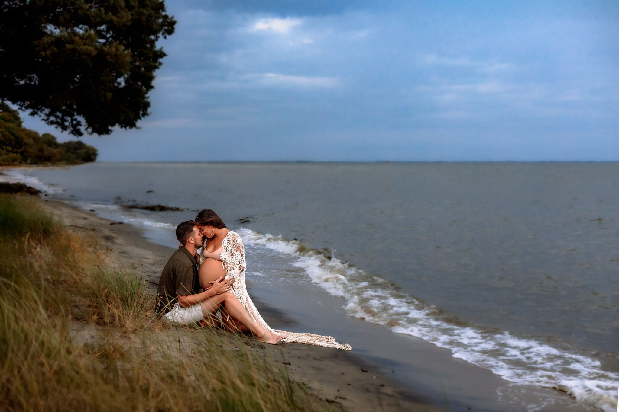 Pregnant couple embracing on the sand with waves behind her during an Annapolis, Maryland beach maternity session.