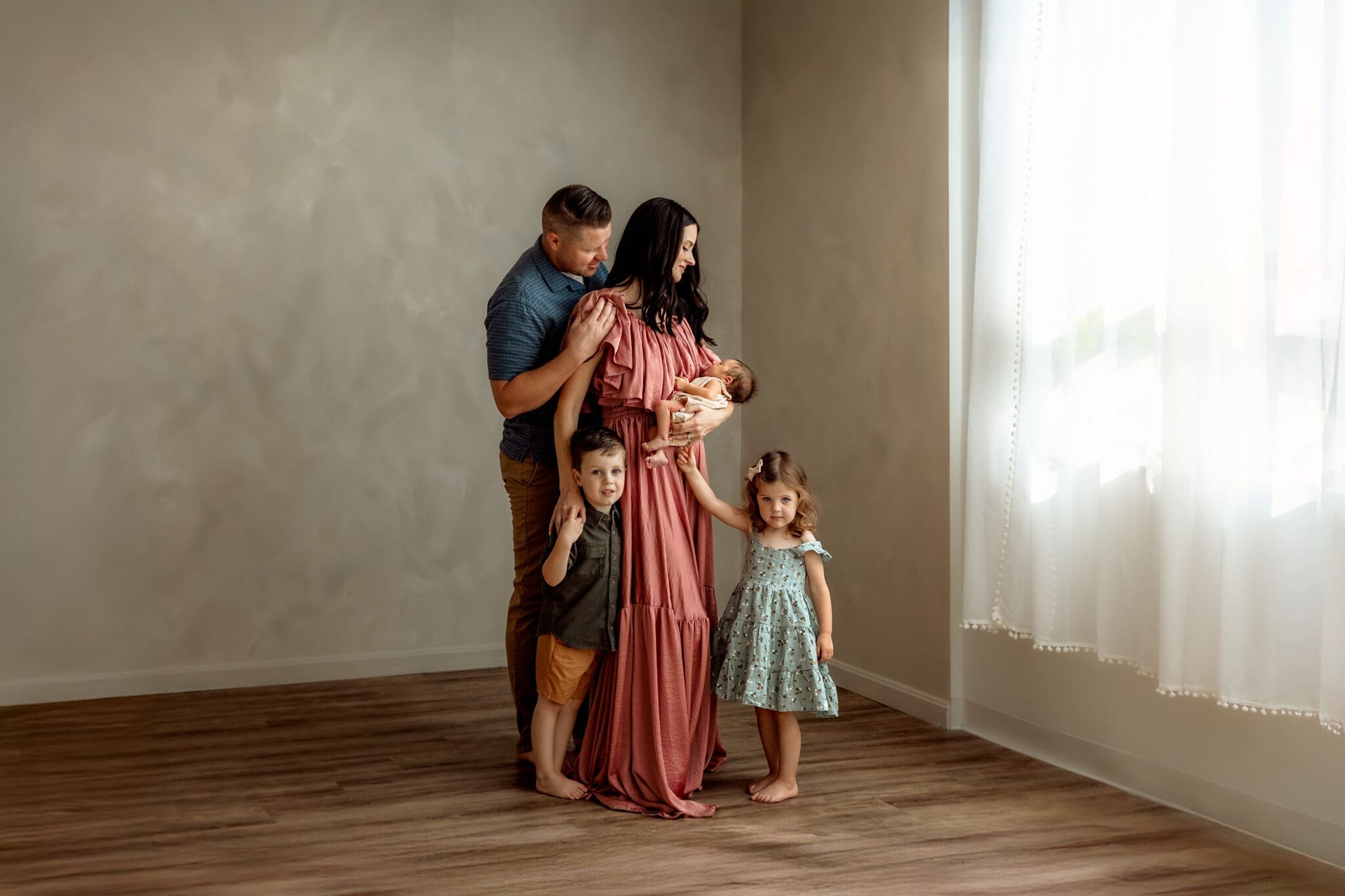 a family in studio. mom holds newborn as she and dad lovingly look down at him. two older siblings stand next to mom and gaze at the camera.