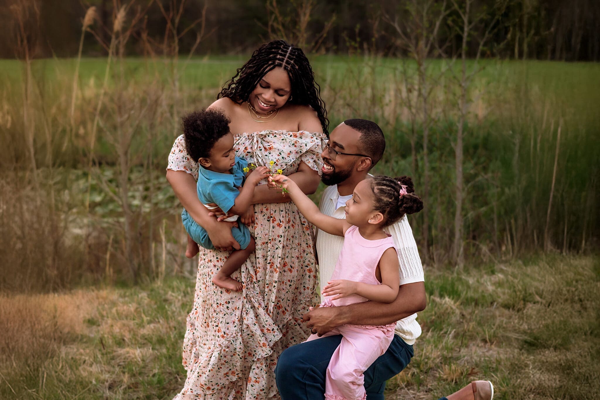 A photo of a smiling family of four sharing a playful moment together in a grassy field, with parents holding their young children on a warm evening. Taken by Erin Link, Maryland Family Photographer in Bowie.